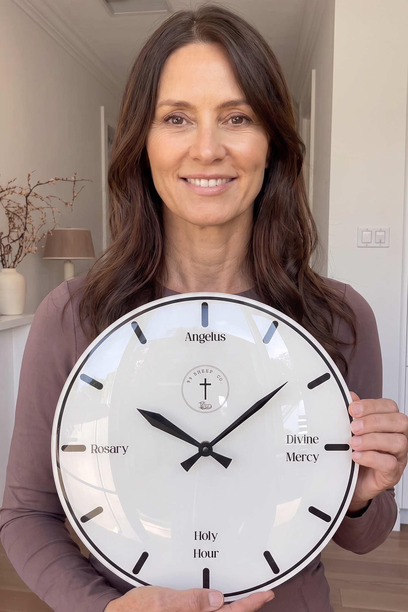 A smiling woman with long brown hair holds a large round white wall clock in a bright, modern hallway. The clock features black hands, black Roman numeral hour markers, and religious text including 'Angels', 'Holy Spirit', 'Divine Mercy', and 'Holy Hour'.
